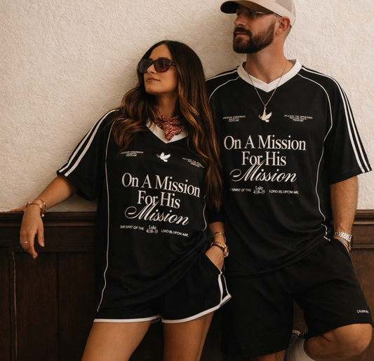 Two people wearing matching black t-shirts with text, standing against a textured wall.