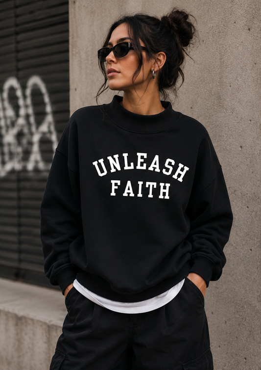 Woman wearing a black 'UNLEASH FAITH' sweatshirt against a concrete wall.