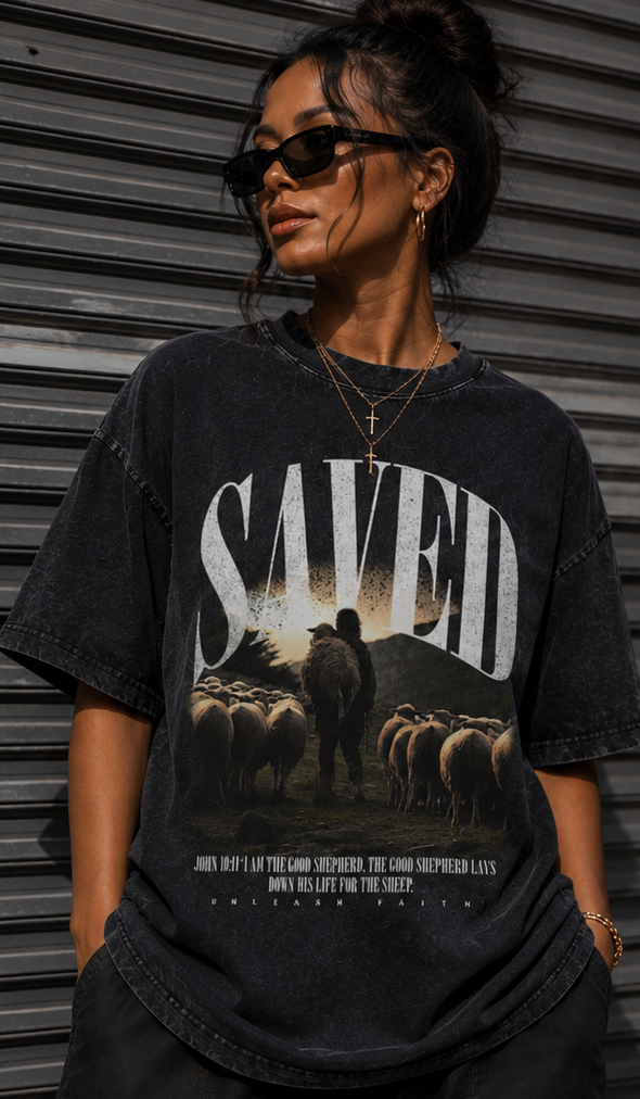 Person wearing a black t-shirt with 'SAVED' graphic design, standing against a metallic wall.